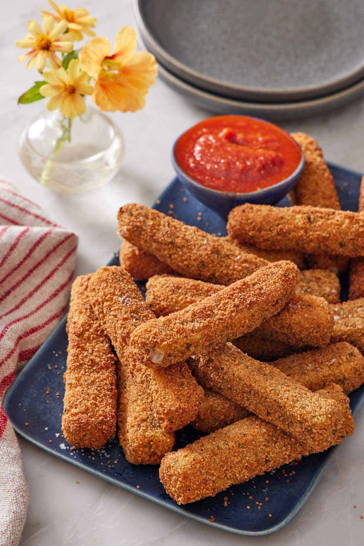 A platter of mozzarella sticks with a bowl of marinara sauce. A stack of plates and vase of flowers in the background.