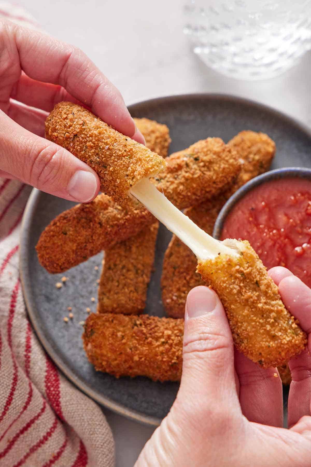 A hand pulling a mozzarella stick in half. A plate of more mozzarella sticks and marinara sauce underneath.