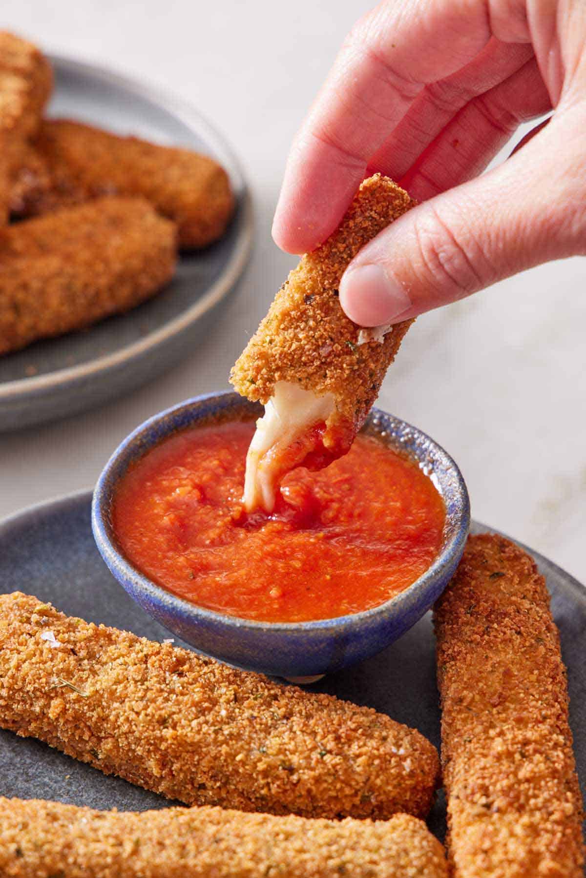 Half of a mozzarella stick dipped into a small bowl of marinara sauce. More mozzarella sticks in the front.