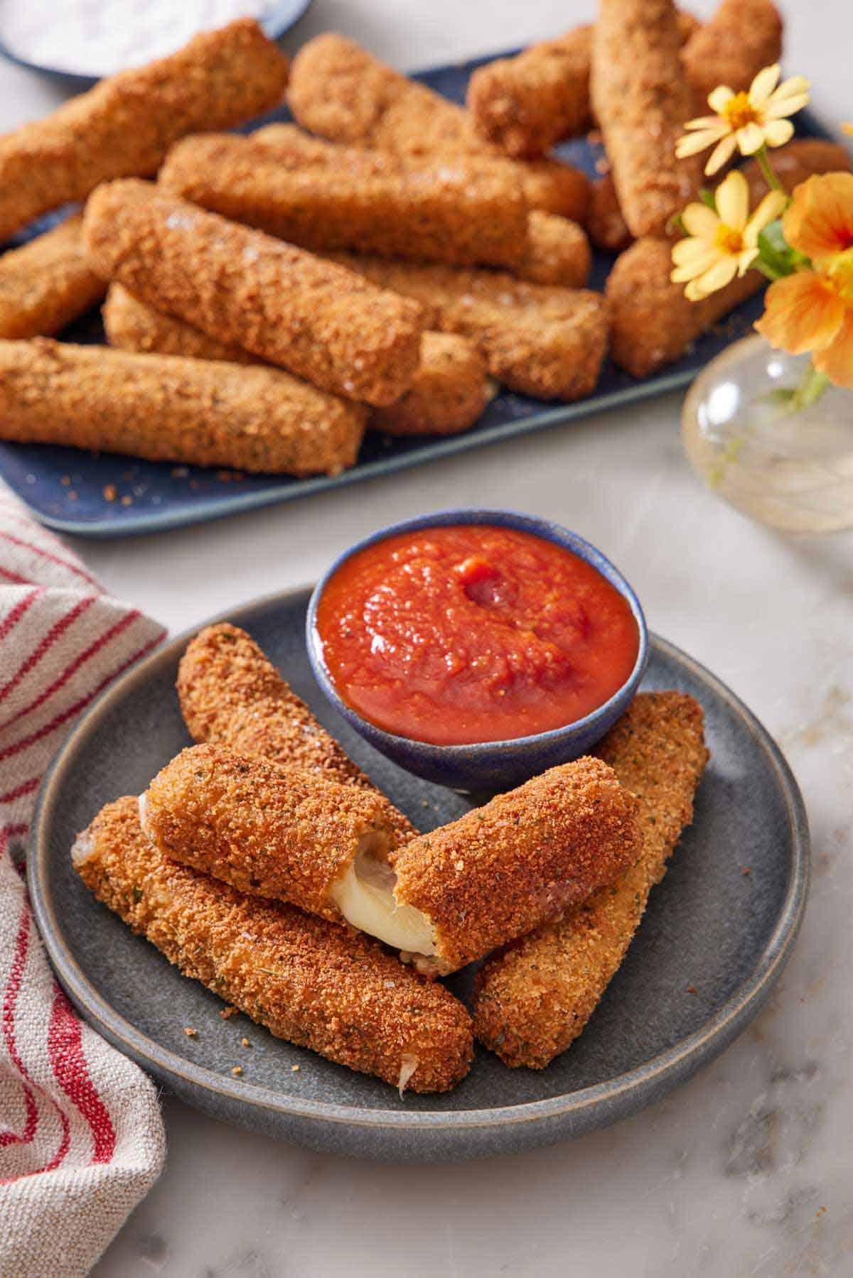 A plate with mozzarella sticks, one torn opened, with a bowl of marinara sauce. A platter of more mozzarella sticks in the background with a small vase of flowers.