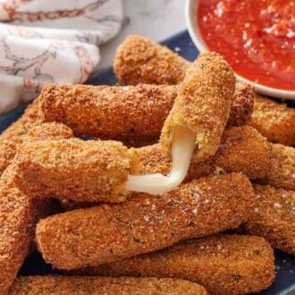 A platter of mozzarella sticks with a bowl of marinara sauce. A torn opened mozzarella stick on top with the cheese coming out.