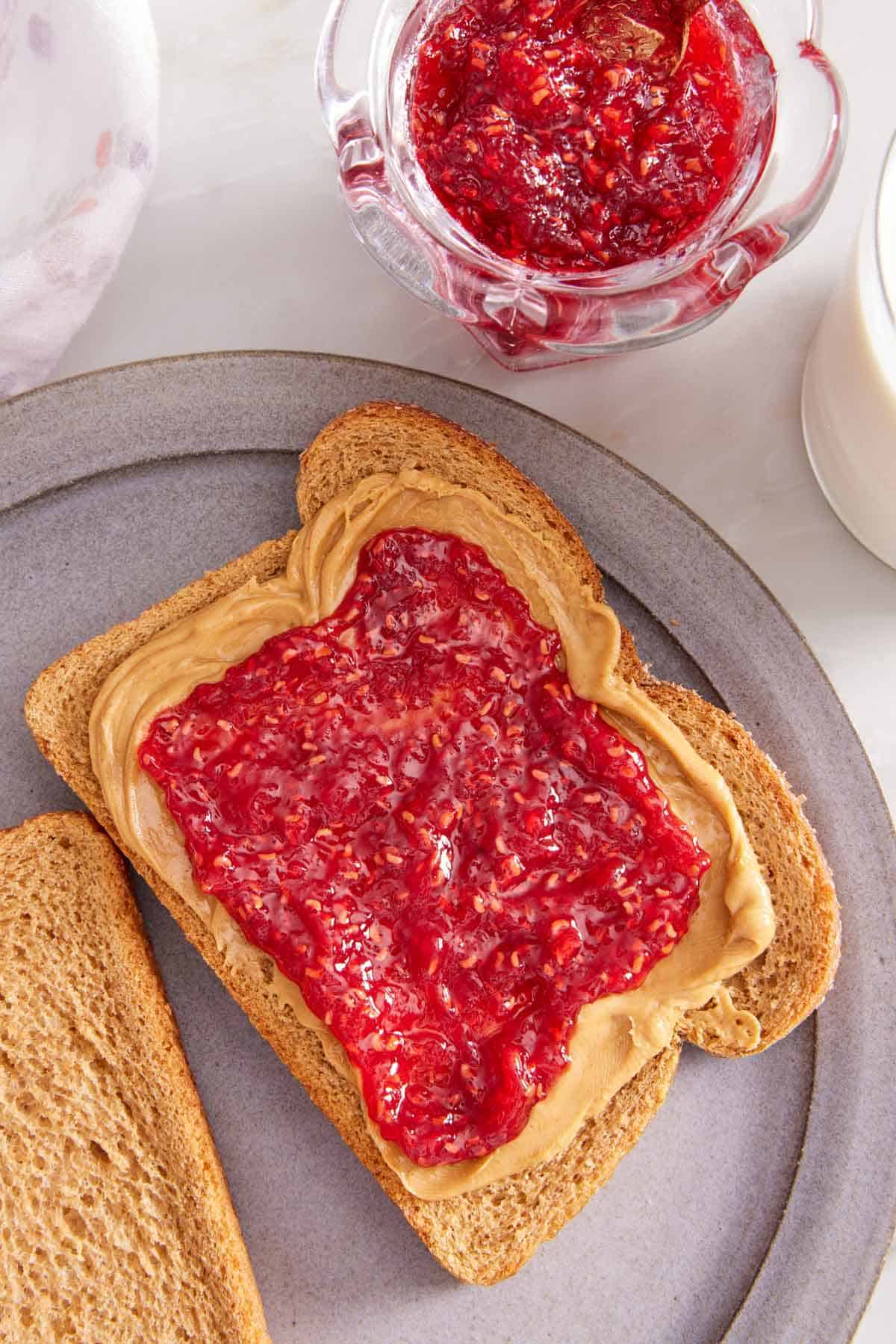 Overhead view of a piece of toast with peanut butter and raspberry jam. A jar of raspberry jam on the side.