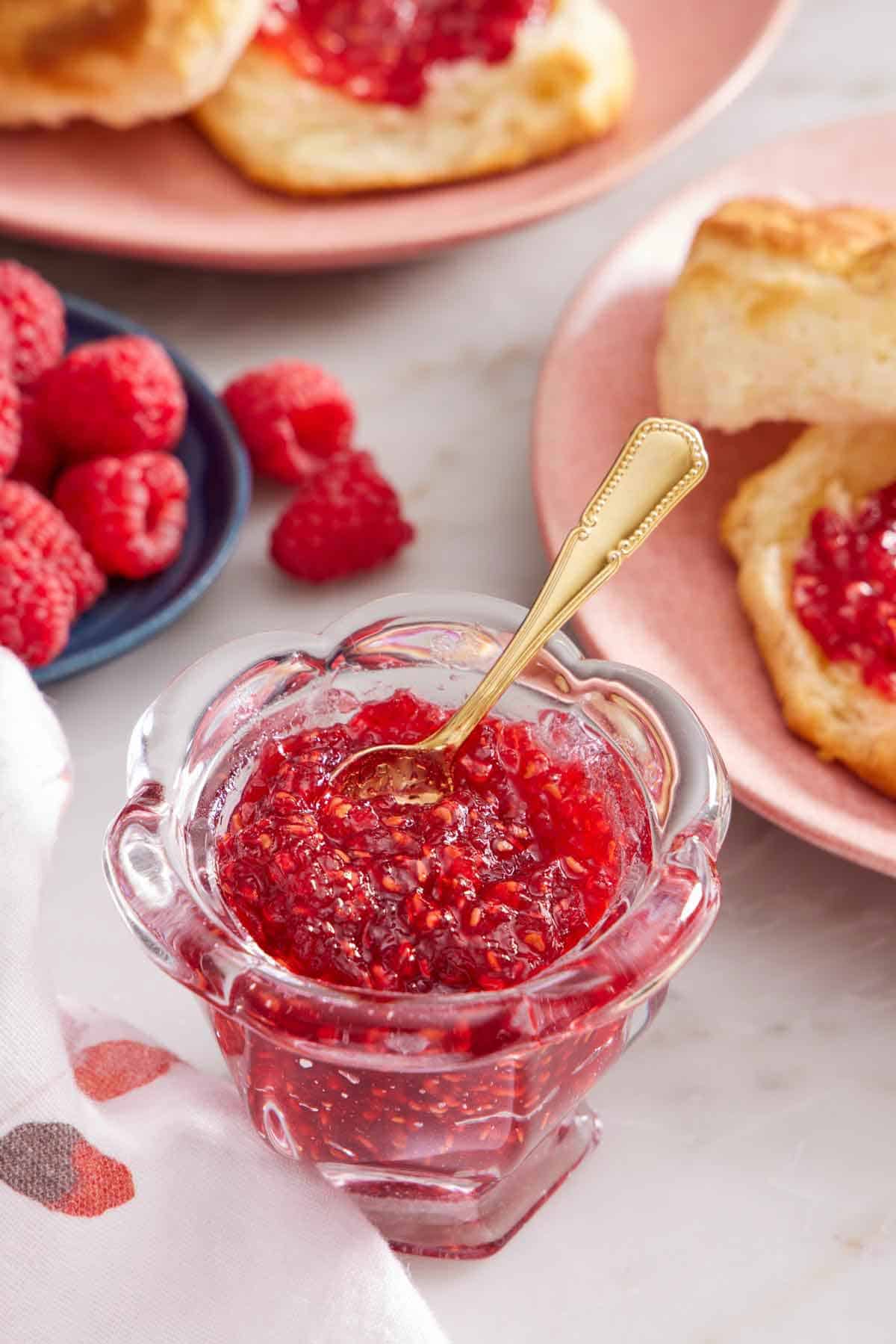 A jar of raspberry jam with a small gold spoon inside. A plate of fresh raspberries in the background along with biscuits with jam on them.