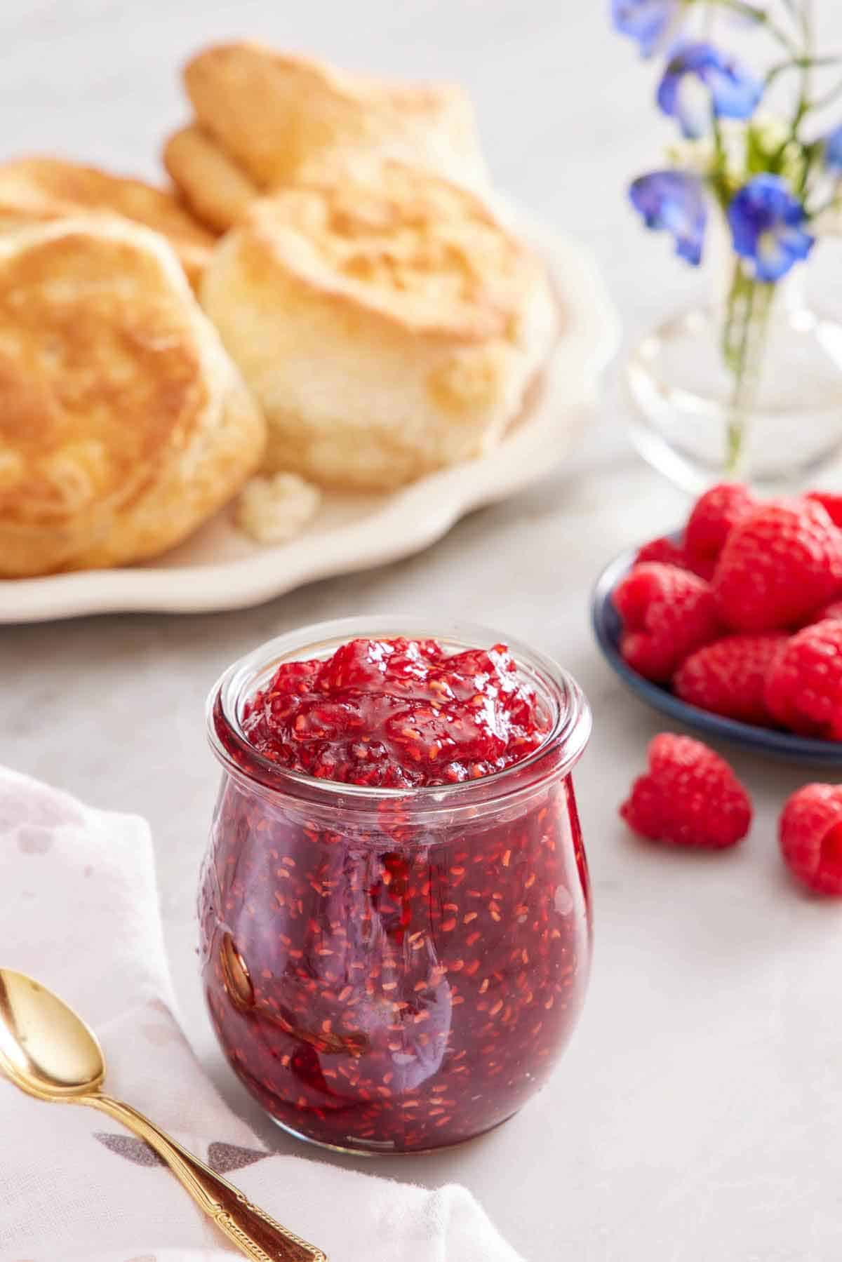 A jar of raspberry jam. A platter of biscuits in the back along with a plate of raspberries and some flowers.
