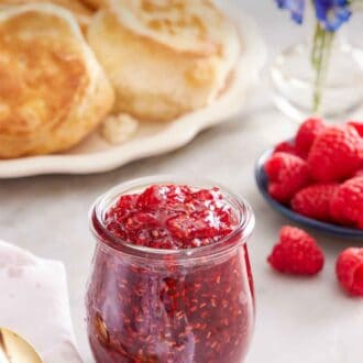 Pinterest graphic of a jar of raspberry jam. A platter of biscuits in the back along with a plate of raspberries and some flowers.