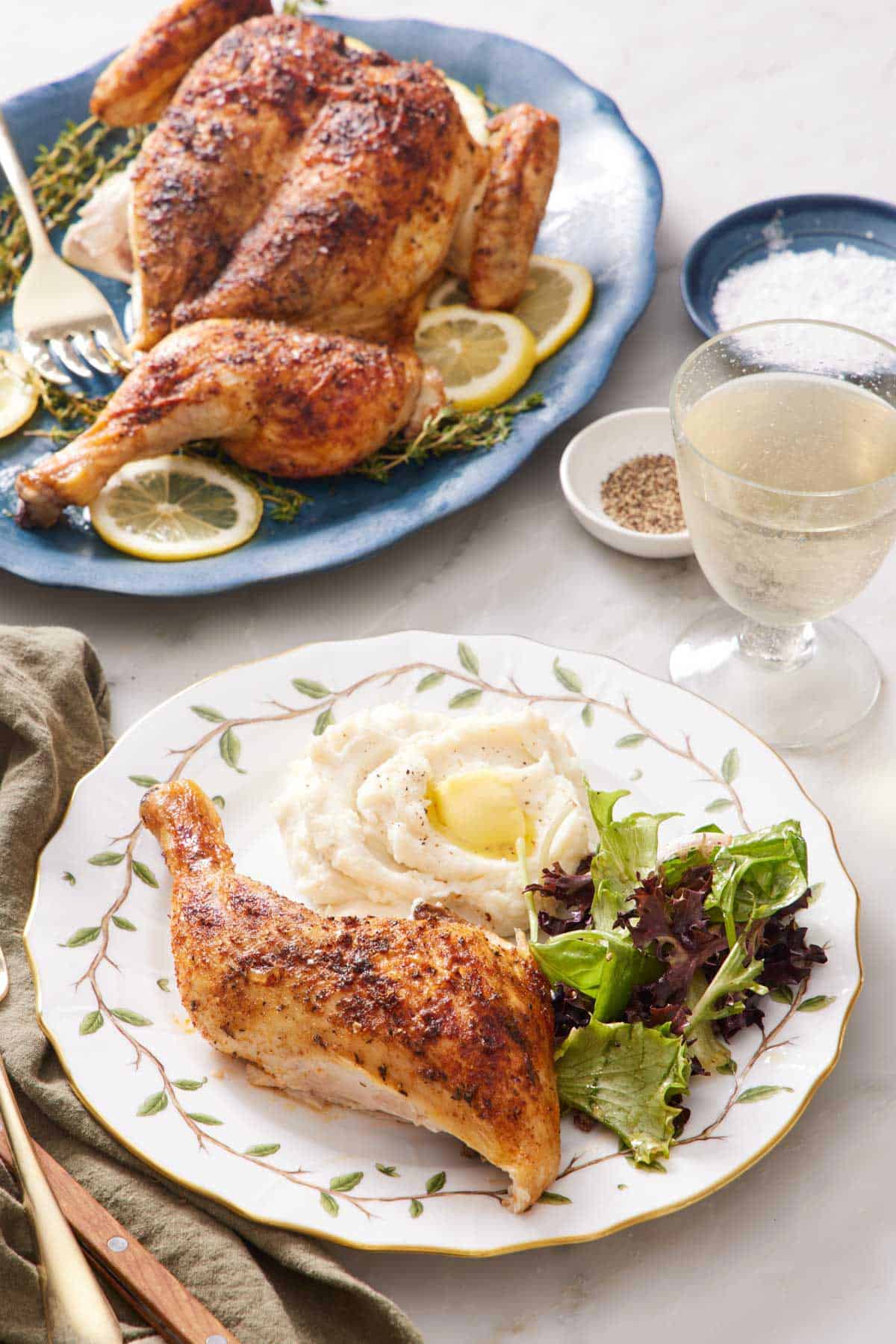 A plate with a chicken leg, mixed greens, and mashed potatoes with the rest of the spatchcock chicken in the background on a platter with a drink, salt, and pepper.