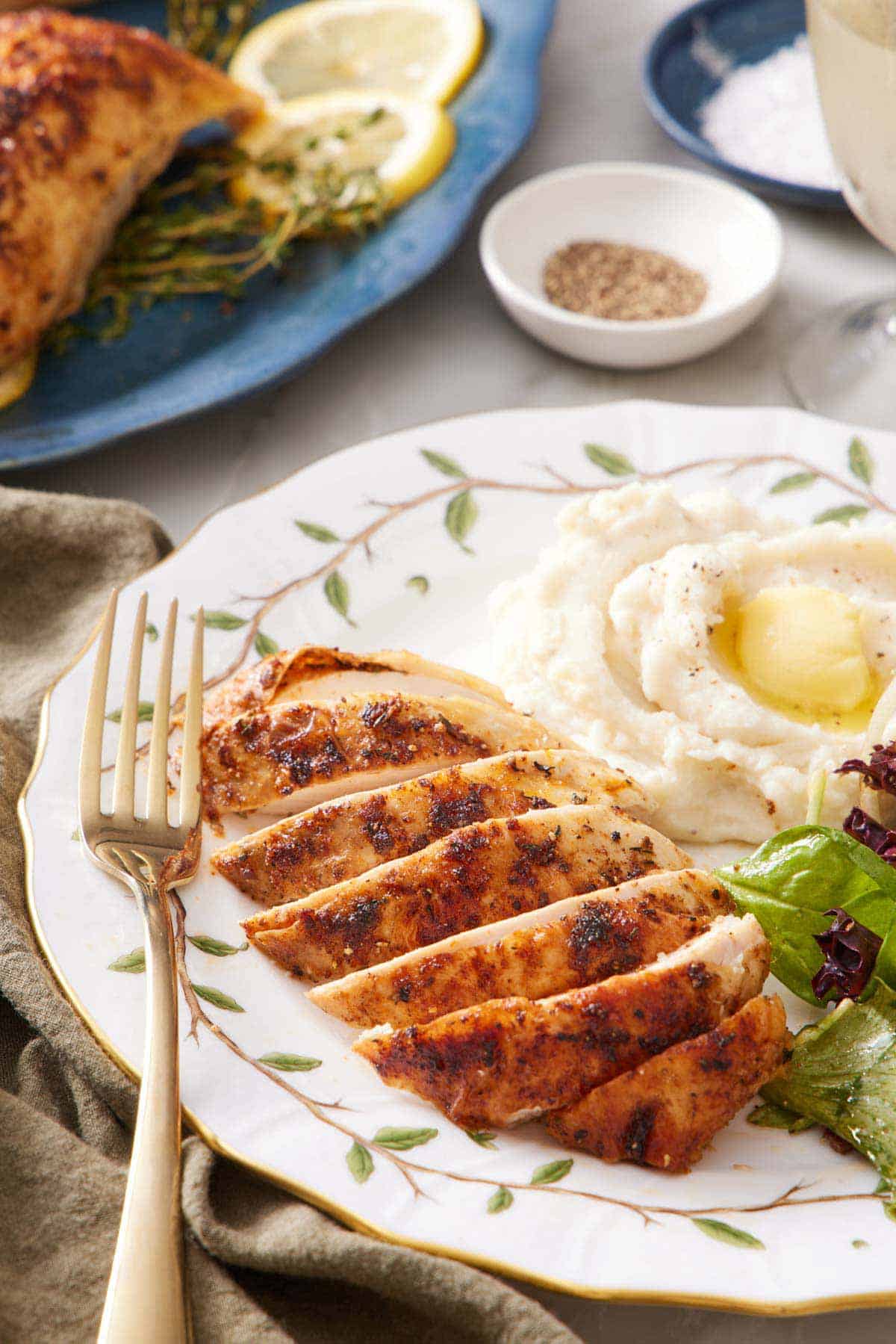 A plate with a spatchcock chicken breast sliced with some mashed potatoes, mixed greens, and a fork.