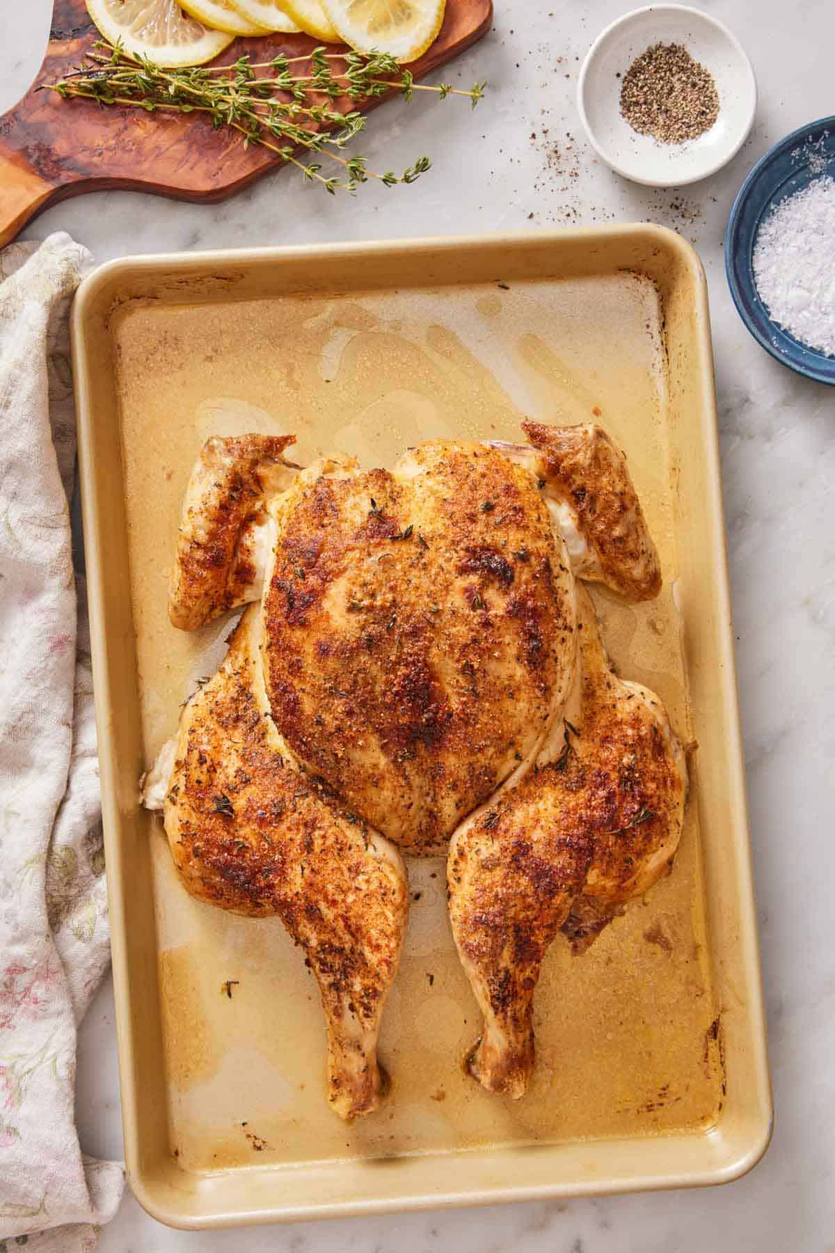 Overhead view of a spatchcock chicken with crispy skin on a sheet pan. Salt, pepper, thyme, and lemon slices off to the side.