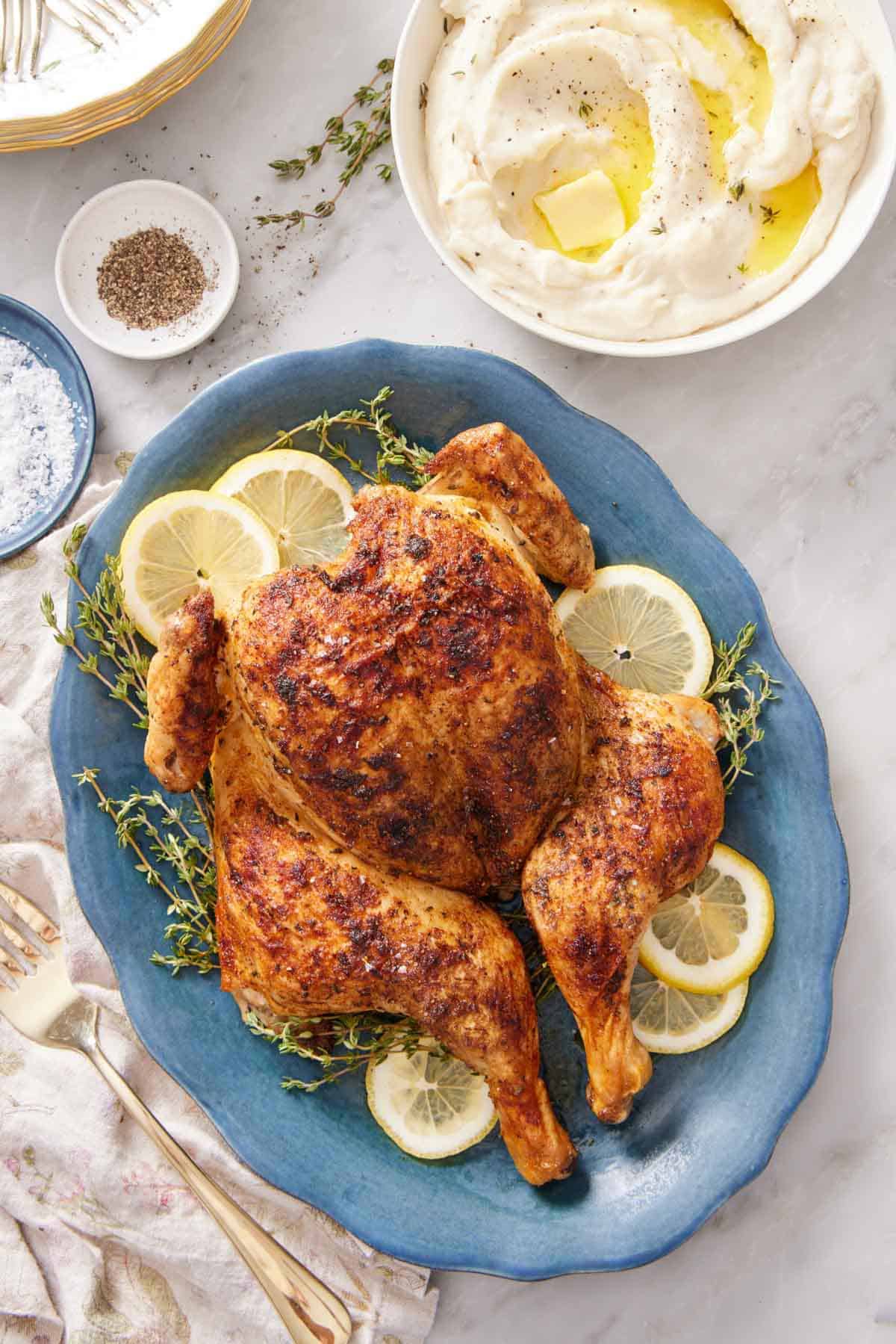 An overhead view of a blue platter of spatchcock chicken with lemon slices and fresh thyme. A bowl of mashed potatoes on the side with salt and pepper.
