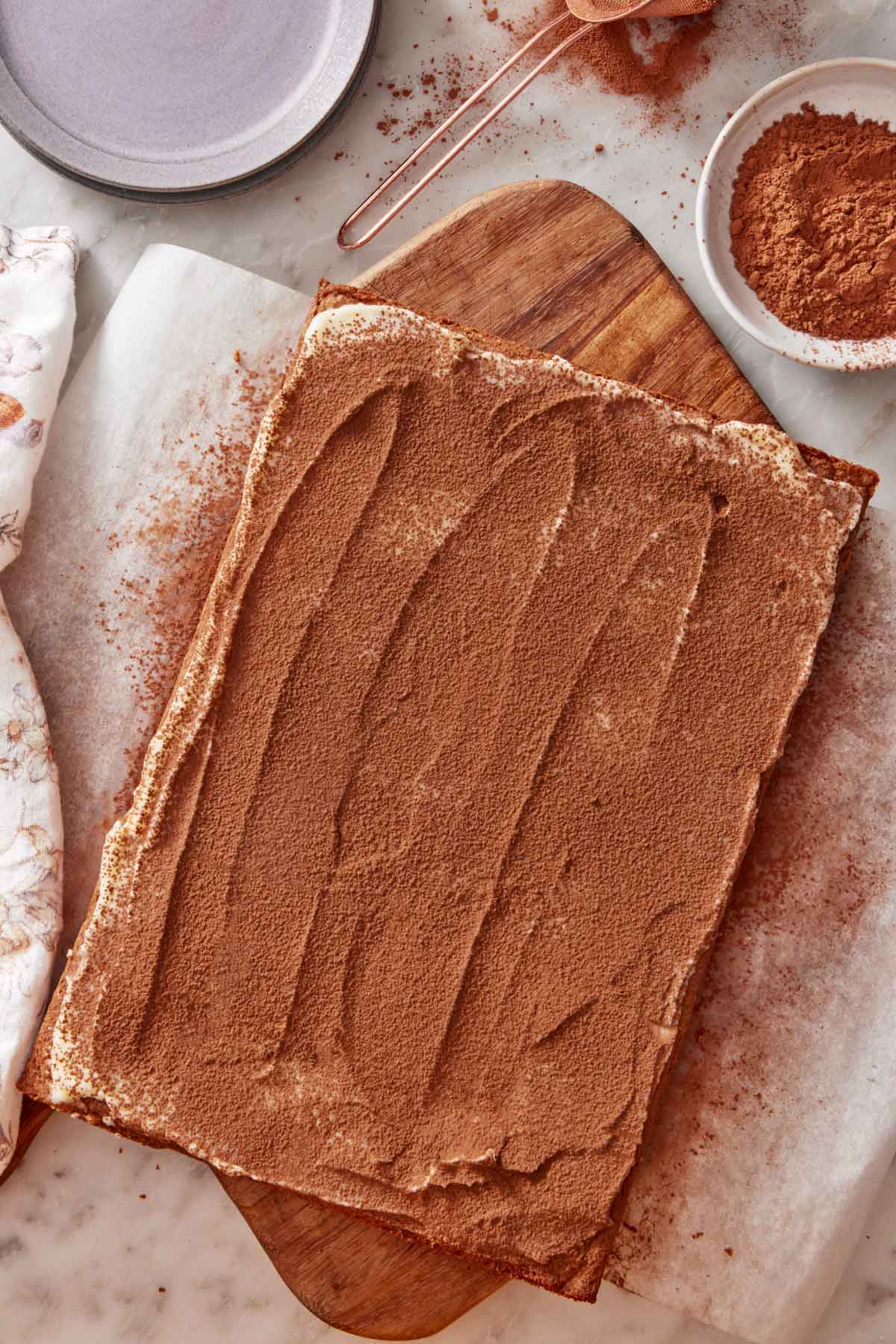 Overhead view of an uncut slab of tiramisu blondies with cocoa powder dusted on top. A bowl of more cocoa powder on the side along with some plates.