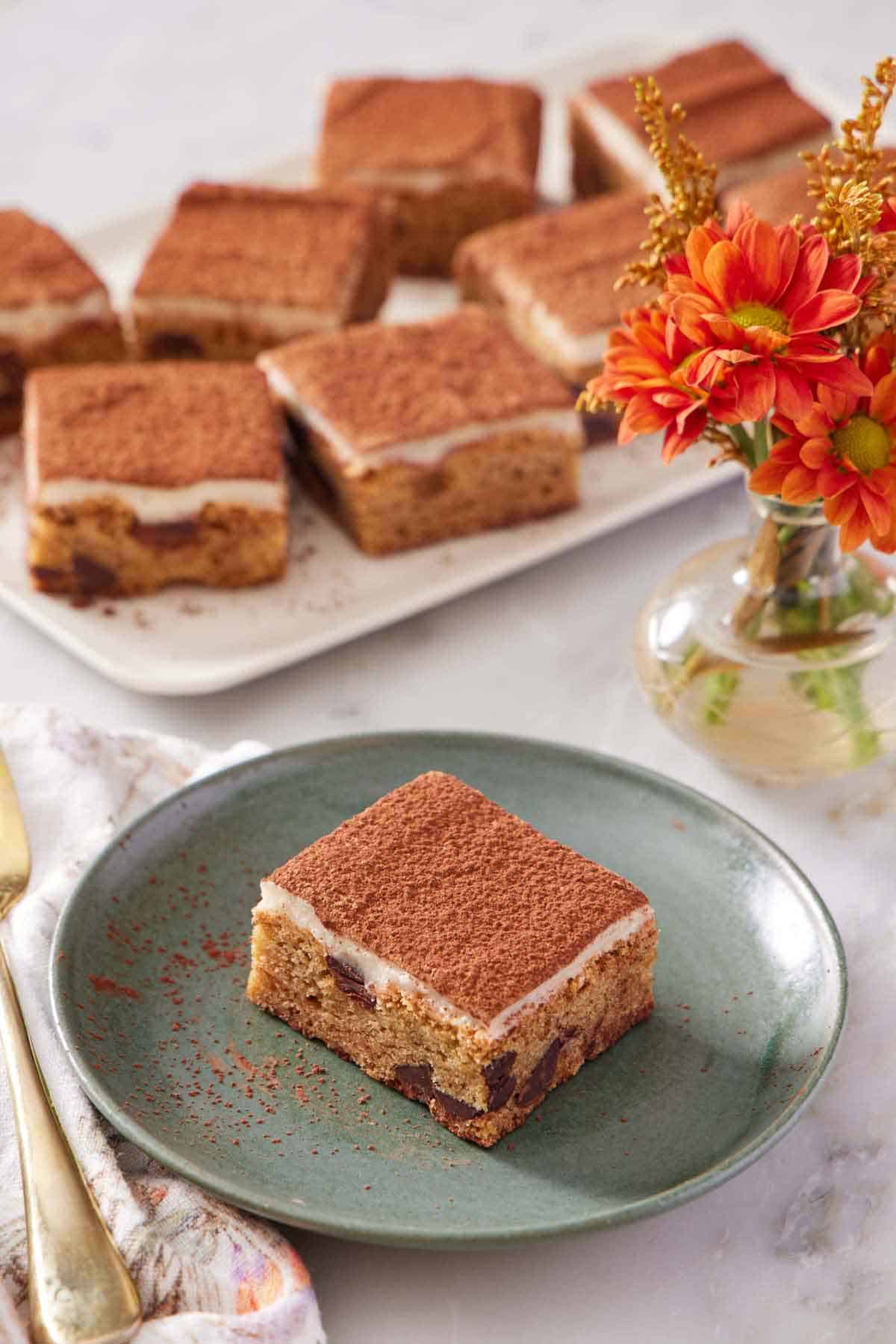 A green plate with a piece of tiramisu blondie. Flowers and a platter of more tiramisu blondies in the background.