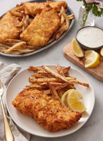 A plate of air fryer fish and chips with lemon wedges. A platter with more in the background also with some lemon wedges and tartar sauce on the side.