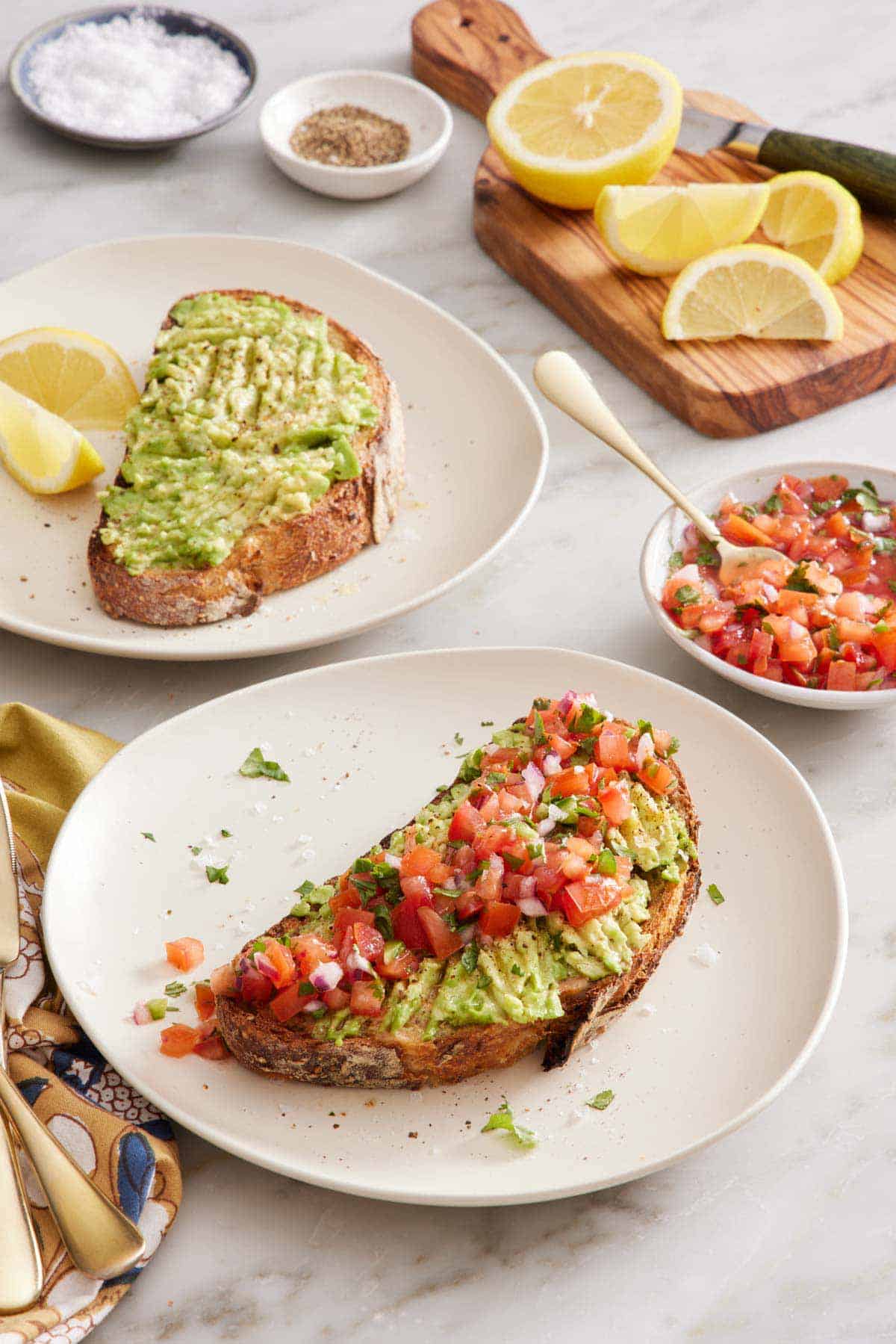 Two plates of avocado toast, one topped with pico de gallo. A bowl of pico de gallo, some cut lemons, and bowls of salt and pepper in the background.