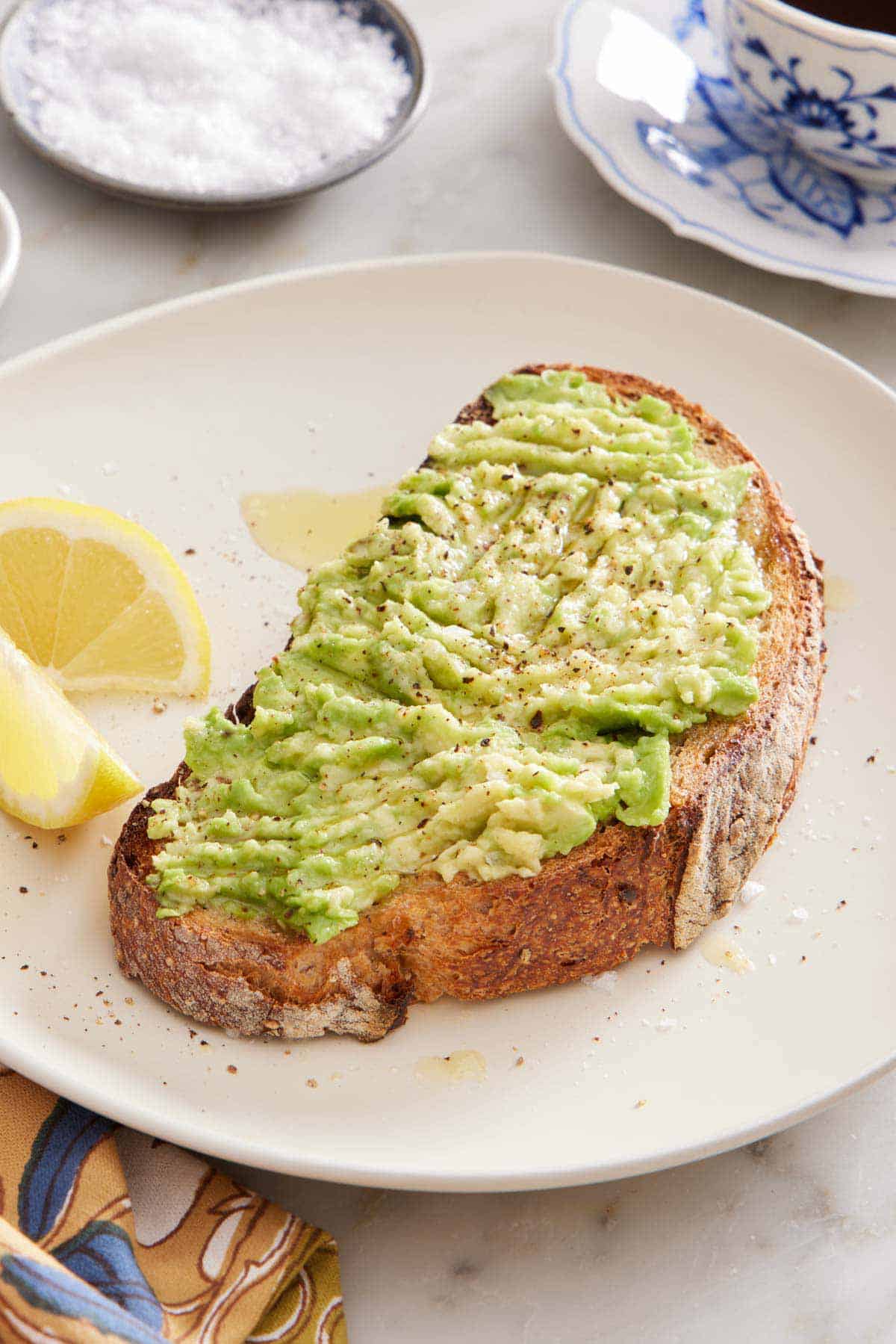 A plate with an avocado toast and lemon wedges. A bowl of salt in the back with a cup of coffee.