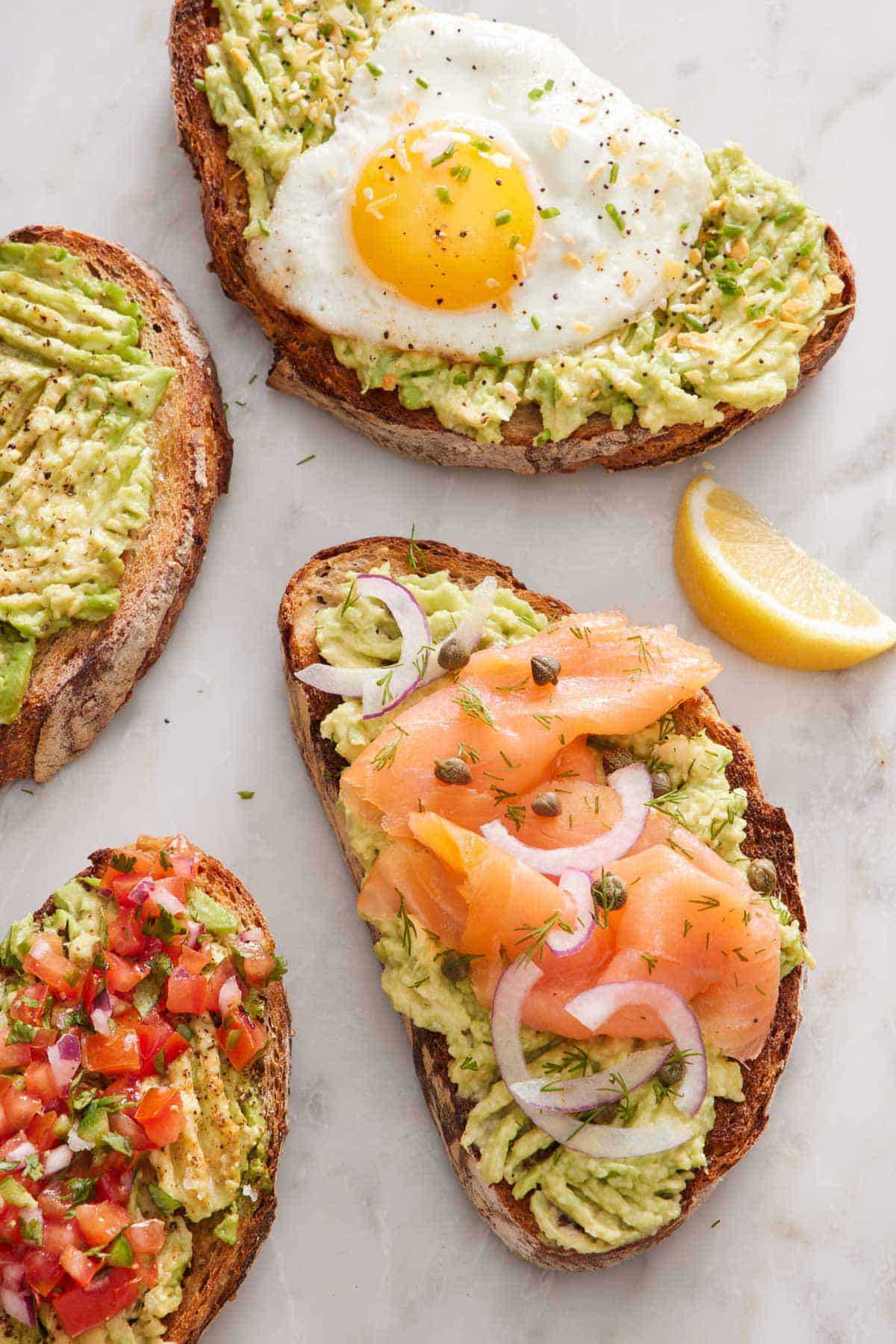 Overhead view of four avocado toasts, one topped with a fried egg, one with smoked salmon, capers, and onions, and one with pico de gallo. A lemon wedge on the side.