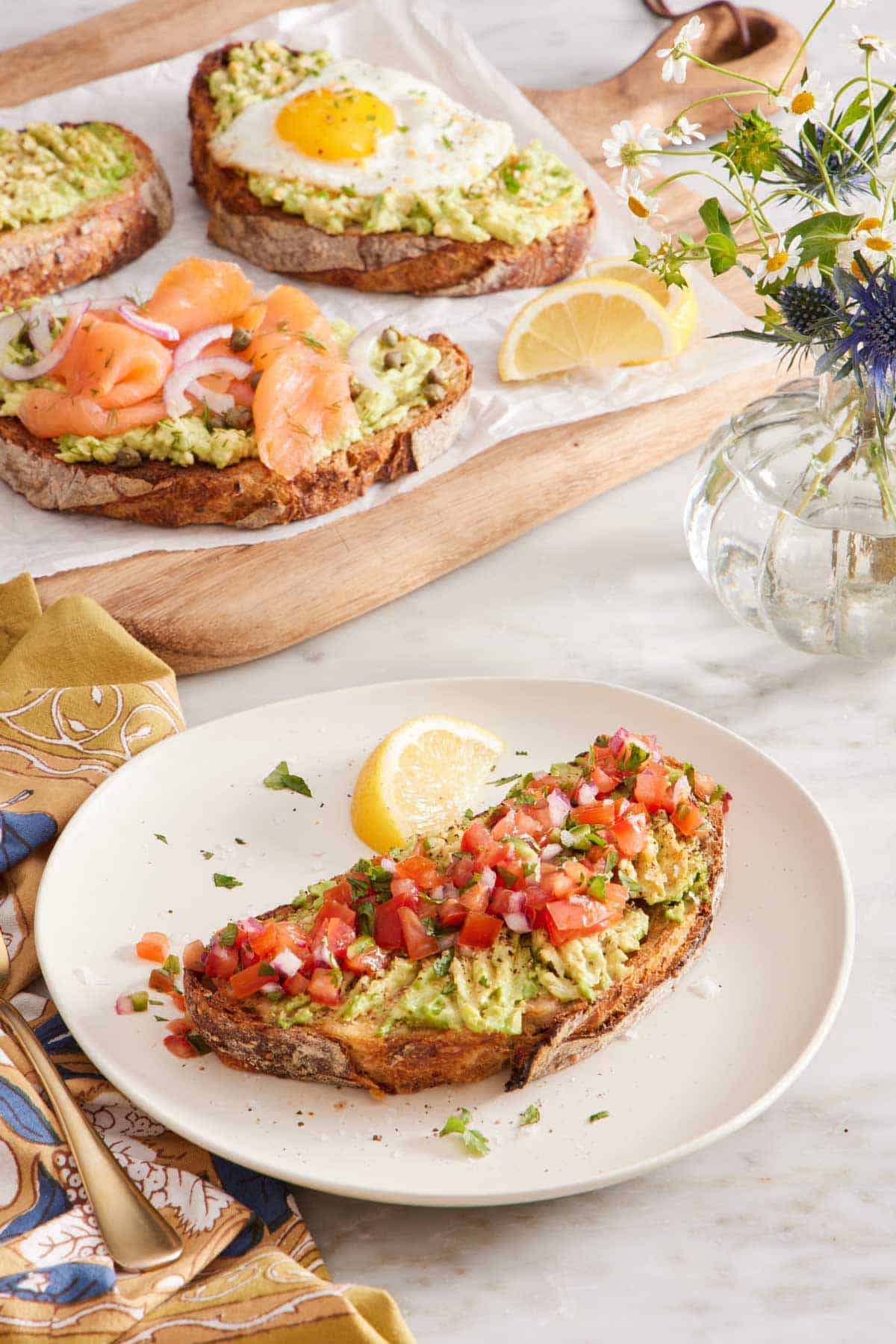 A plate with avocado toast topped with pico de gallo and a lemon wedge on the side. A wooden serving board in the background with three pieces of avocado toast with various toppings and some flowers on the side.