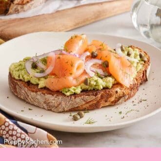 Pinterest graphic of avocado toast topped with smoked salmon, capers, and onions on a plate. More avocado toast in the back.