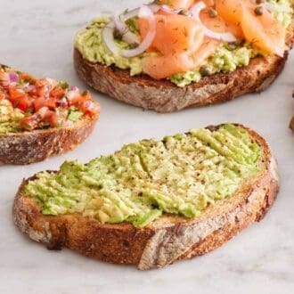 Four avocado toasts, one topped with a fried egg, one with smoked salmon, capers, and onions, and one with pico de gallo.