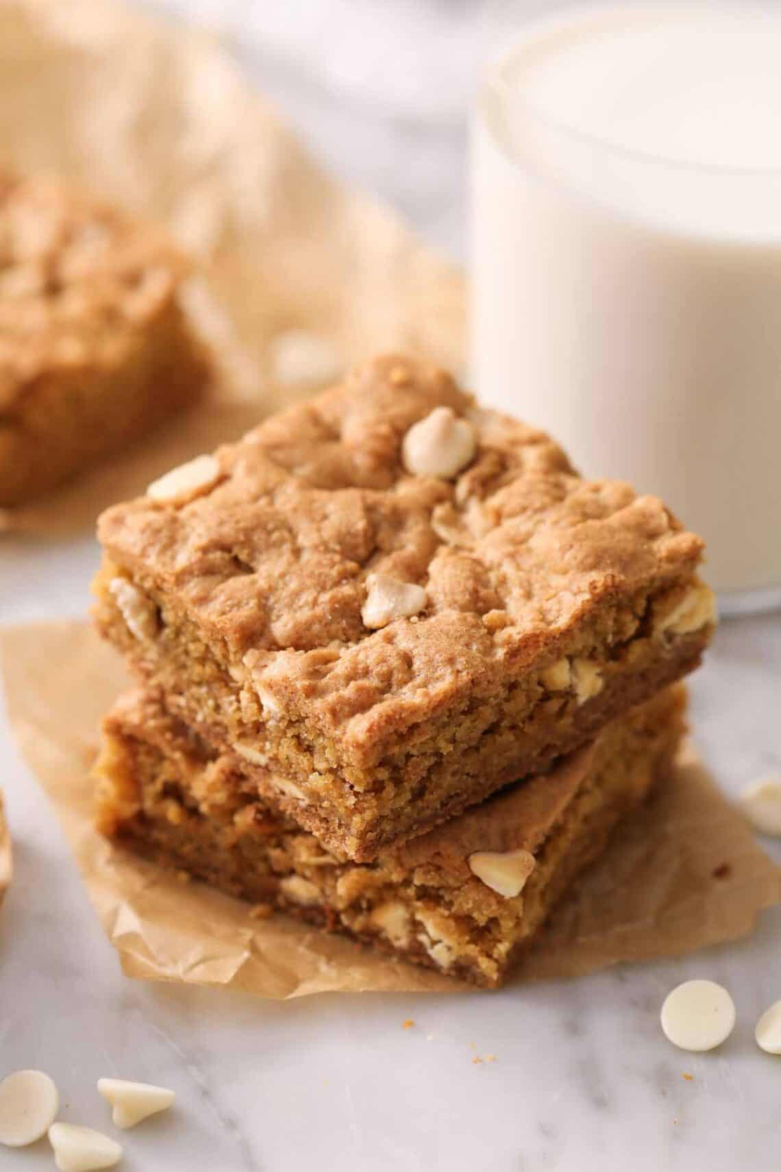 Two blondies, stacked, on a piece of brown parchment paper. A glass of milk in the background.