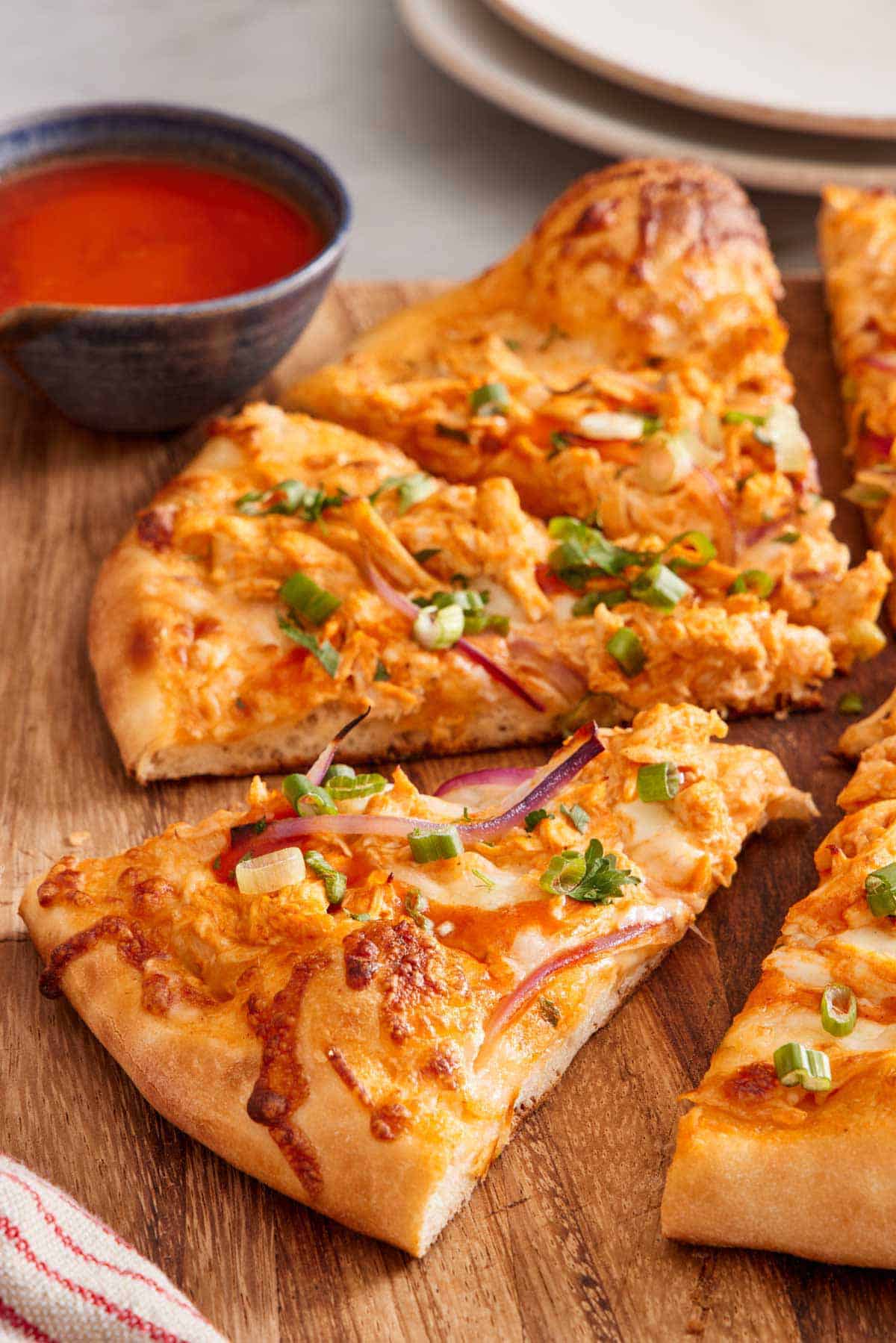 Buffalo chicken pizza cut on a wooden serving board with a slice slightly pulled forward. Buffalo sauce in the background.