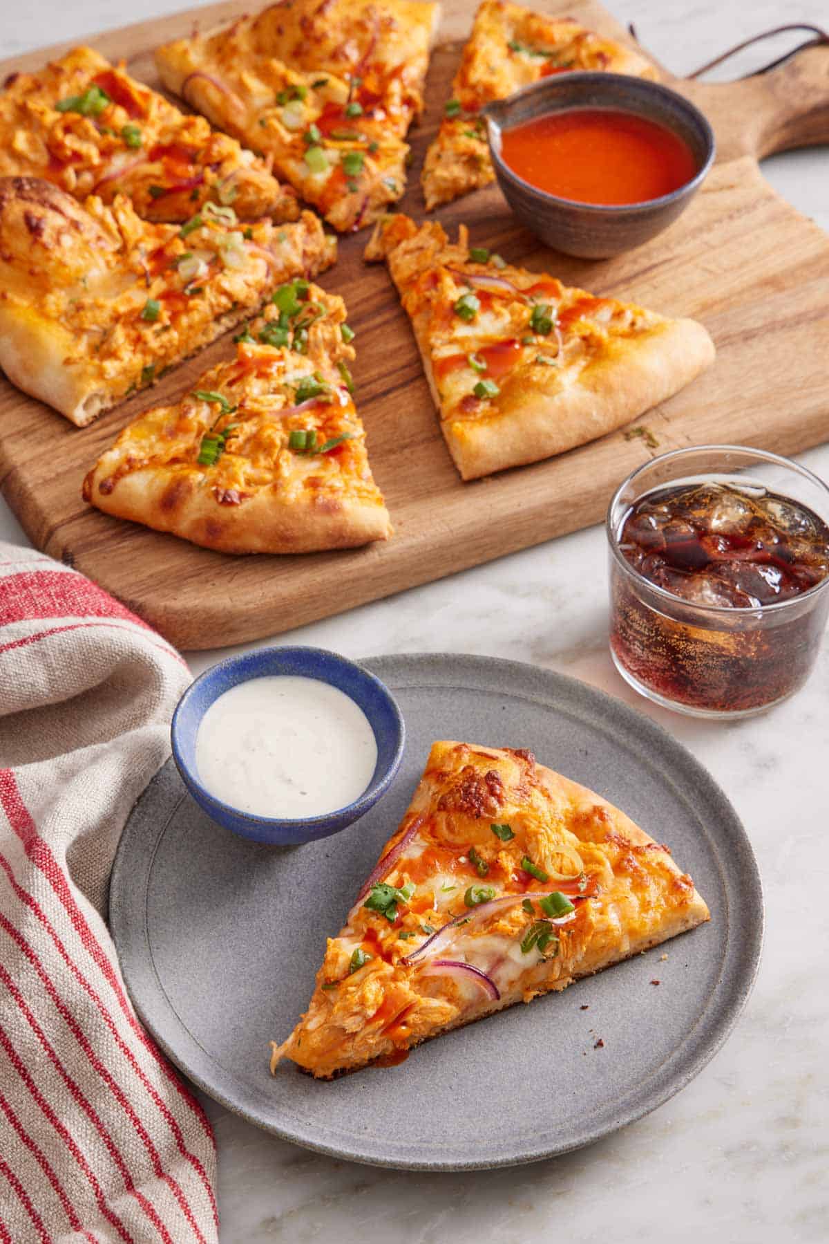 A plate with a slice of buffalo chicken pizza with a bowl of dip. The rest of the pizza in the background with more buffalo sauce on the side and a glass of soda.