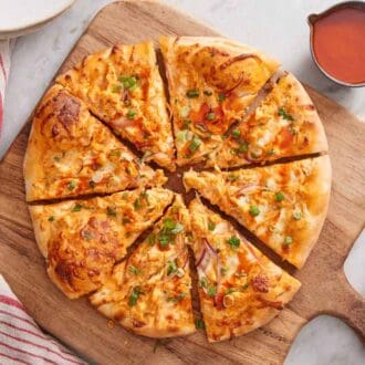 Overhead view of a sliced buffalo chicken pizza on a wooden serving board. A bowl of buffalo sauce on the side with a pizza cutter.