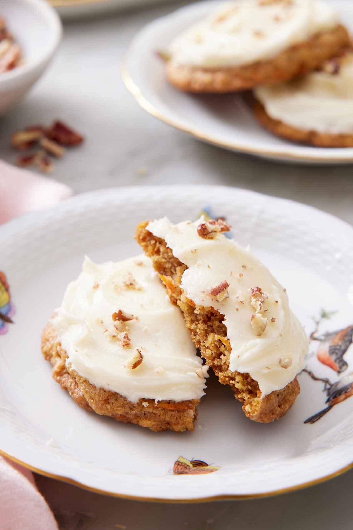 A plate with a carrot cake cookie broken in half. Another plate with two cookies in the background.