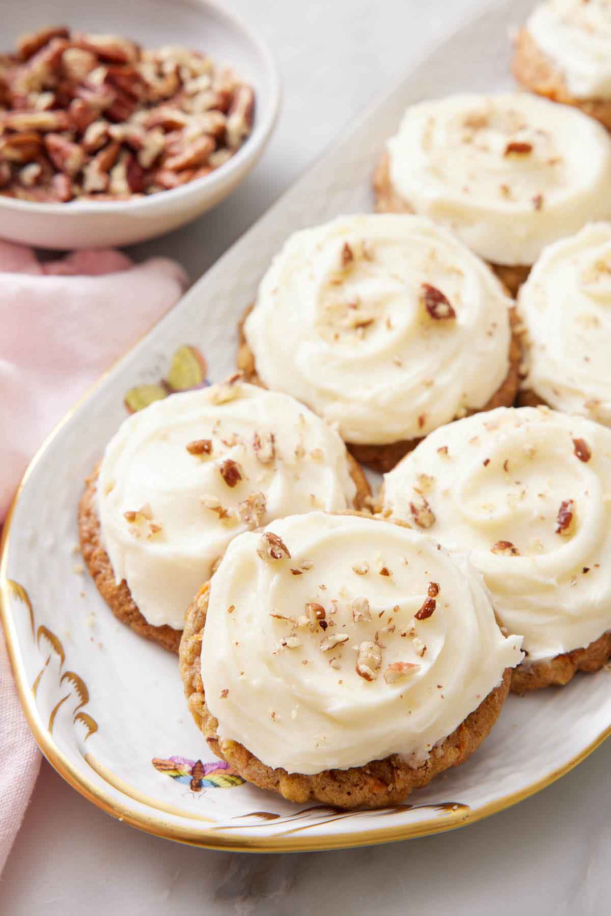 A platter with multiple carrot cake cookies with cream cheese topping. A bowl of pecans in the background.