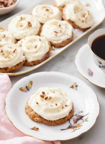 A plate with a carrot cake cookie with a platter in the background with more cookies along with a cup of coffee.