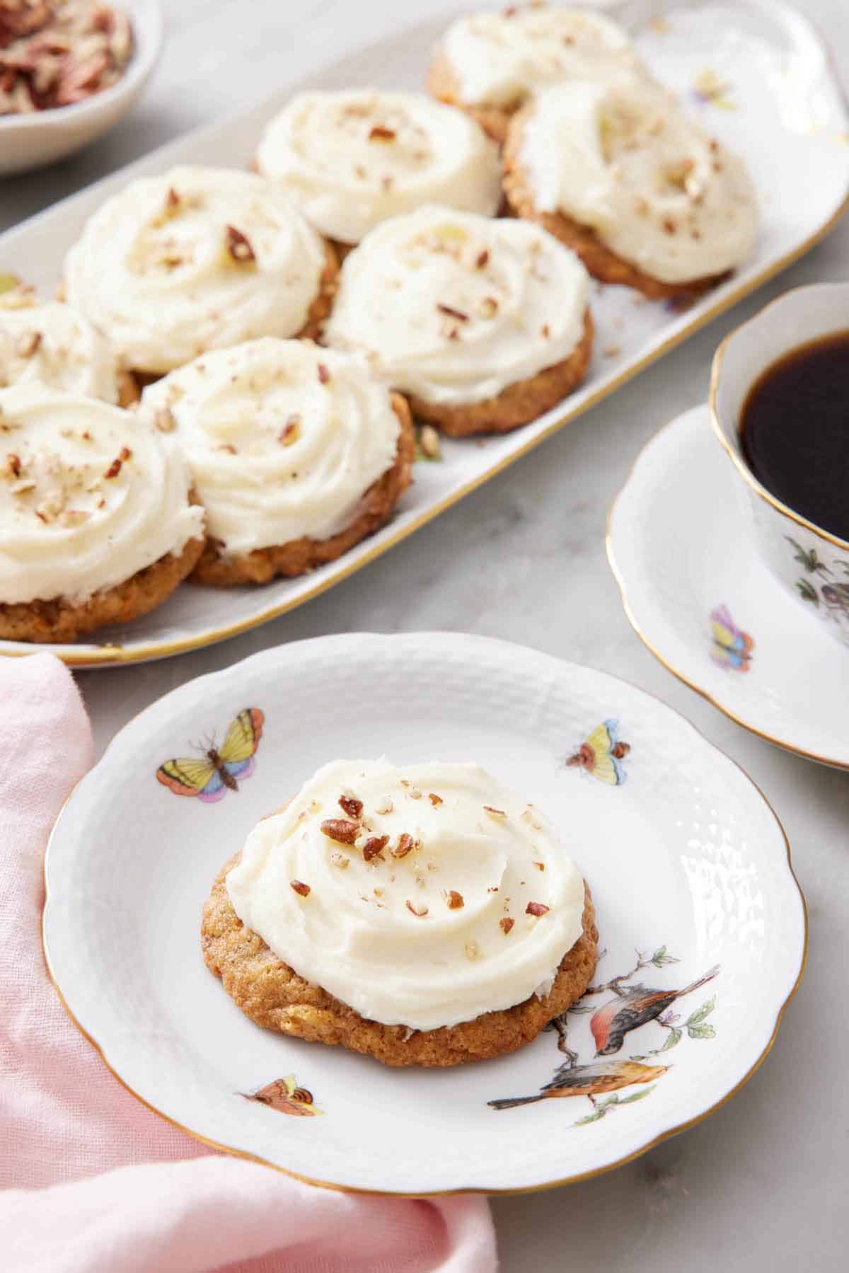A plate with a carrot cake cookie with a platter in the background with more cookies along with a cup of coffee.