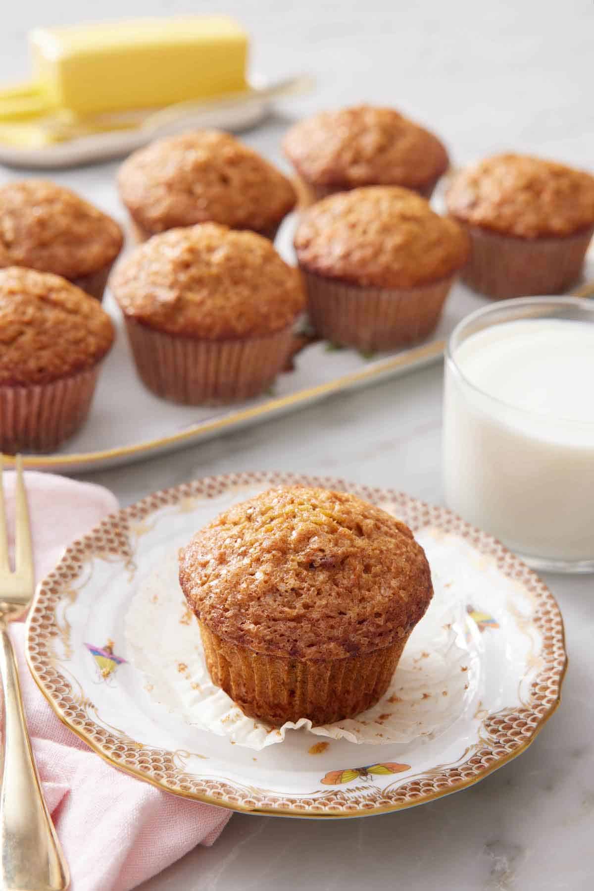 A plate with a carrot muffin with the liner pulled down. A glass of milk in the background with a platter of more muffins.