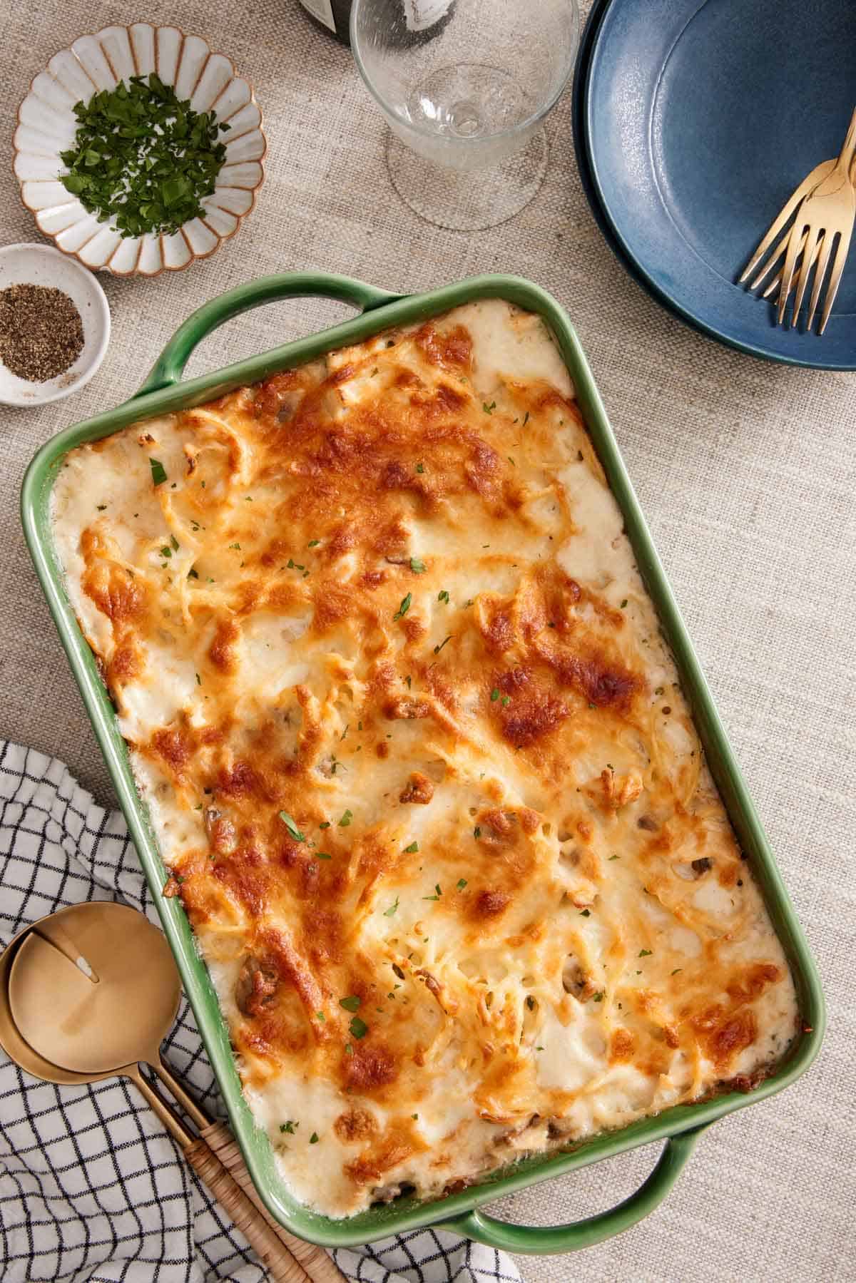 Overhead view of a green baking dish of chicken tetrazzini. A bowl of chopped parsley and pepper on the side along with serving spoons, plates, and forks.