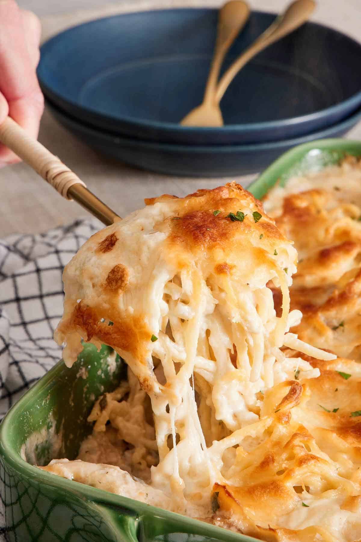 A serving of chicken tetrazzini lifted from a green baking dish. Bowls and forks in the background.