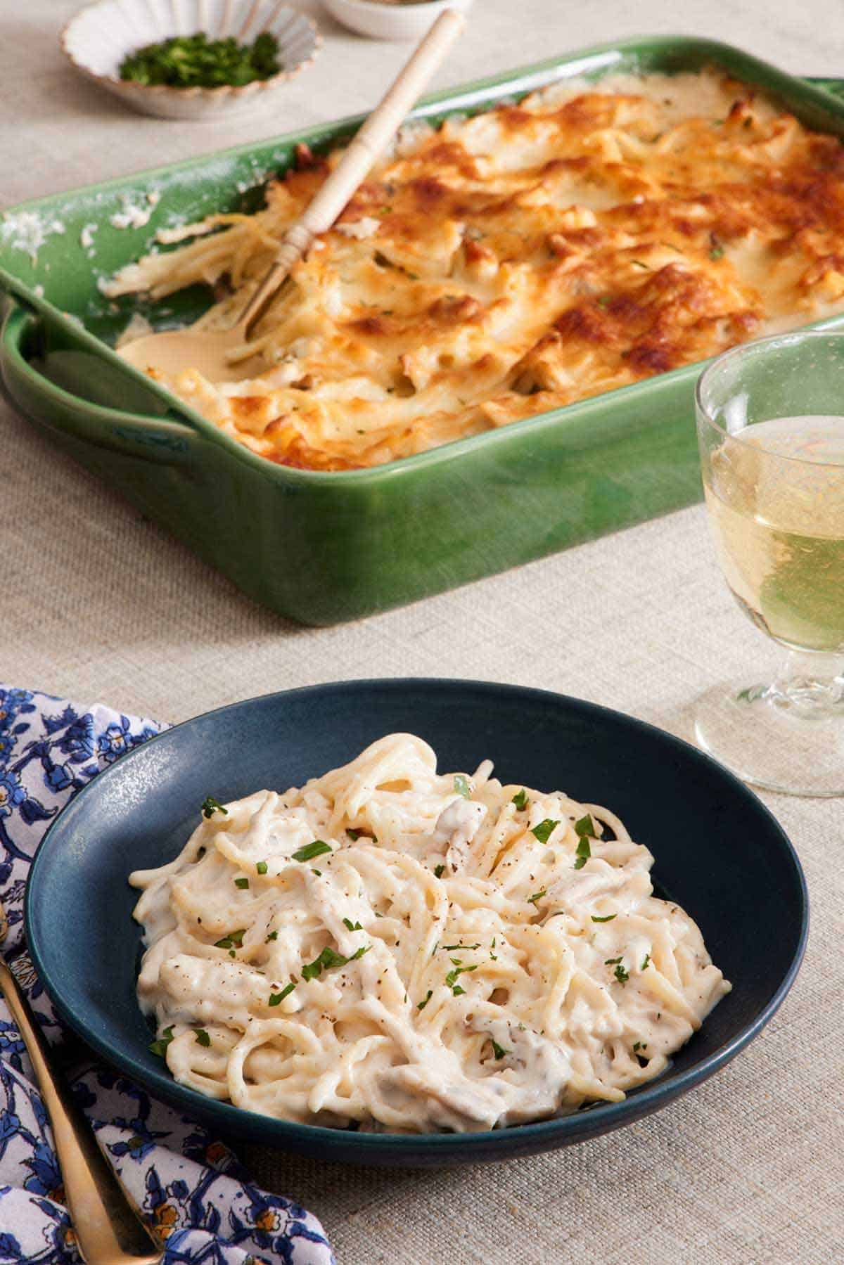 A bowl of chicken tetrazzini with the baking dish of more chicken tetrazzini in the background along with a drink.