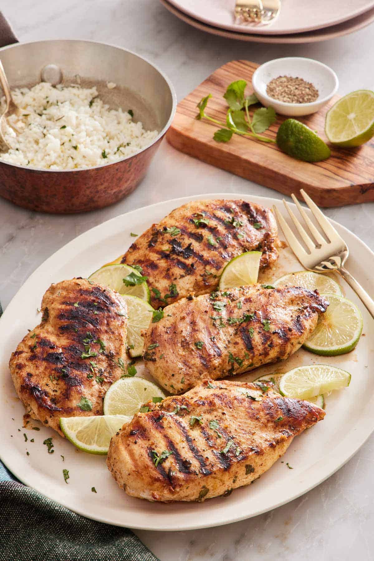 A platter with four cilantro lime chicken along with cut limes and a fork. A pot of cilantro lime rice in the background along with garnishes.