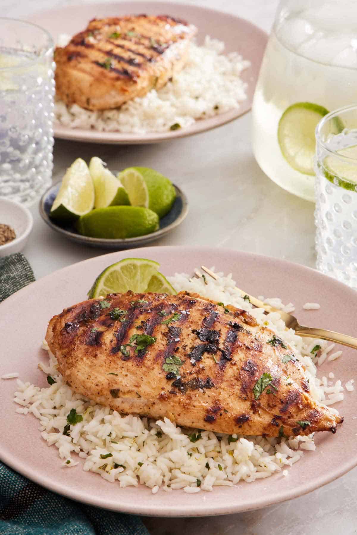 A plate with a cilantro lime chicken on top of a bed of rice with a fork. A bowl of lime wedges, another plated serving, and drinks in the background.