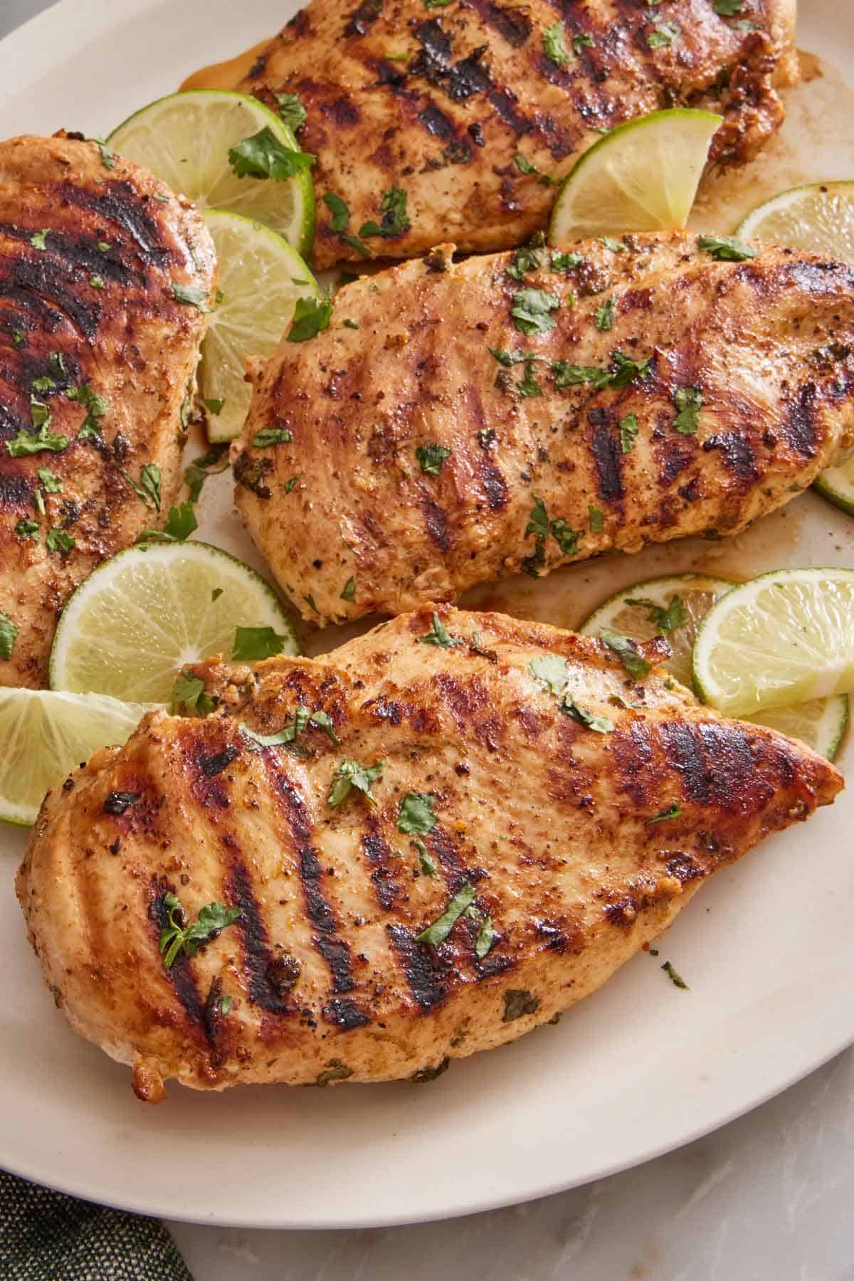 A close up view of a cilantro lime chicken breast on a platter with more chicken along with cut limes and cilantro garnish.