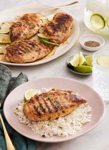 A plate with a cilantro lime chicken breast over a bed of rice and a lime wedge. A platter with more chicken in the background with limes and a drink.