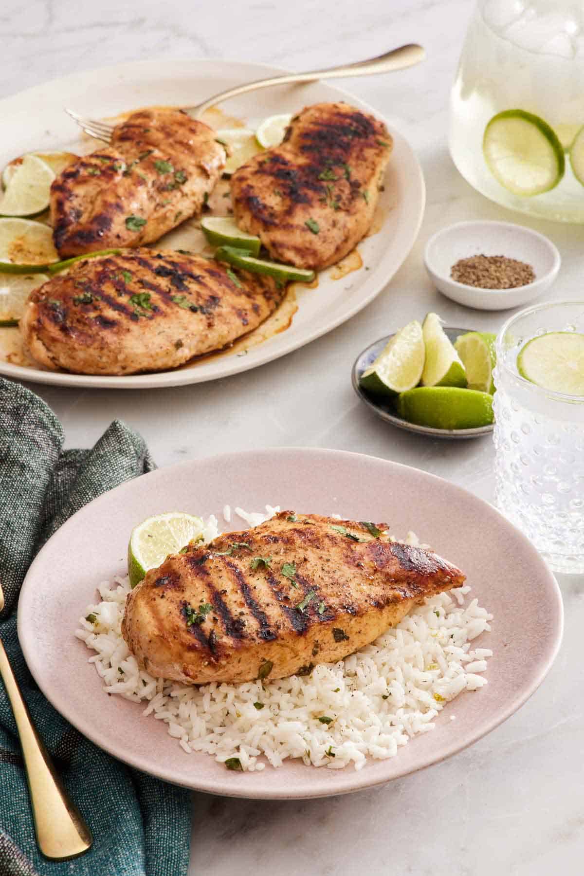 A plate with a cilantro lime chicken breast over a bed of rice and a lime wedge. A platter with more chicken in the background with limes and a drink.