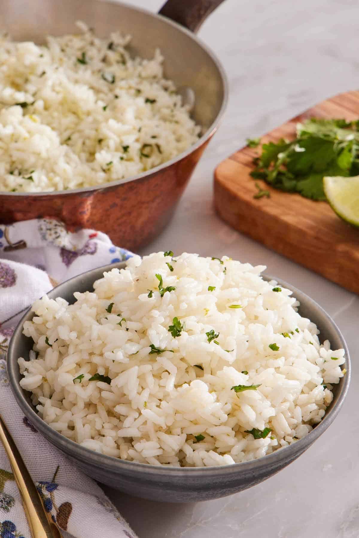 A bowl of cilantro lime rice with chopped cilantro on top. A pot of more cilantro lime rice in the background along with some cut lime and cilantro.