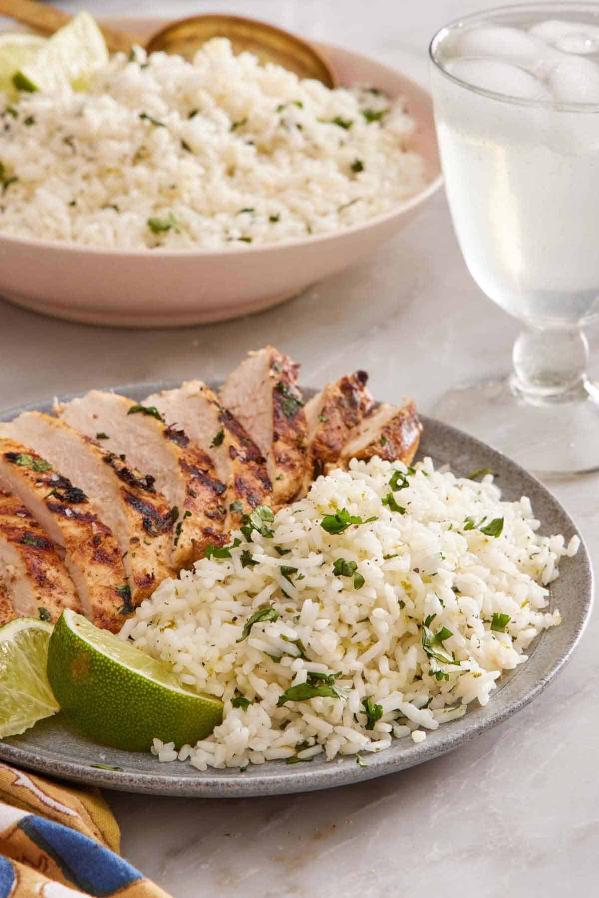 A plate of cilantro lime rice with sliced chicken breast and cut limes. A drink in the background along with more rice.