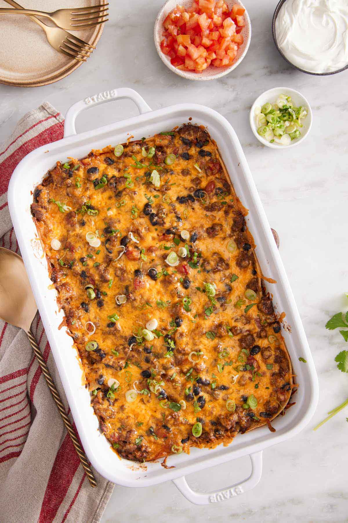 Overhead view of a baking dish of Mexican lasagna with green onion garnish. Bowls of garnishes on the side along with a stack of plates and forks.