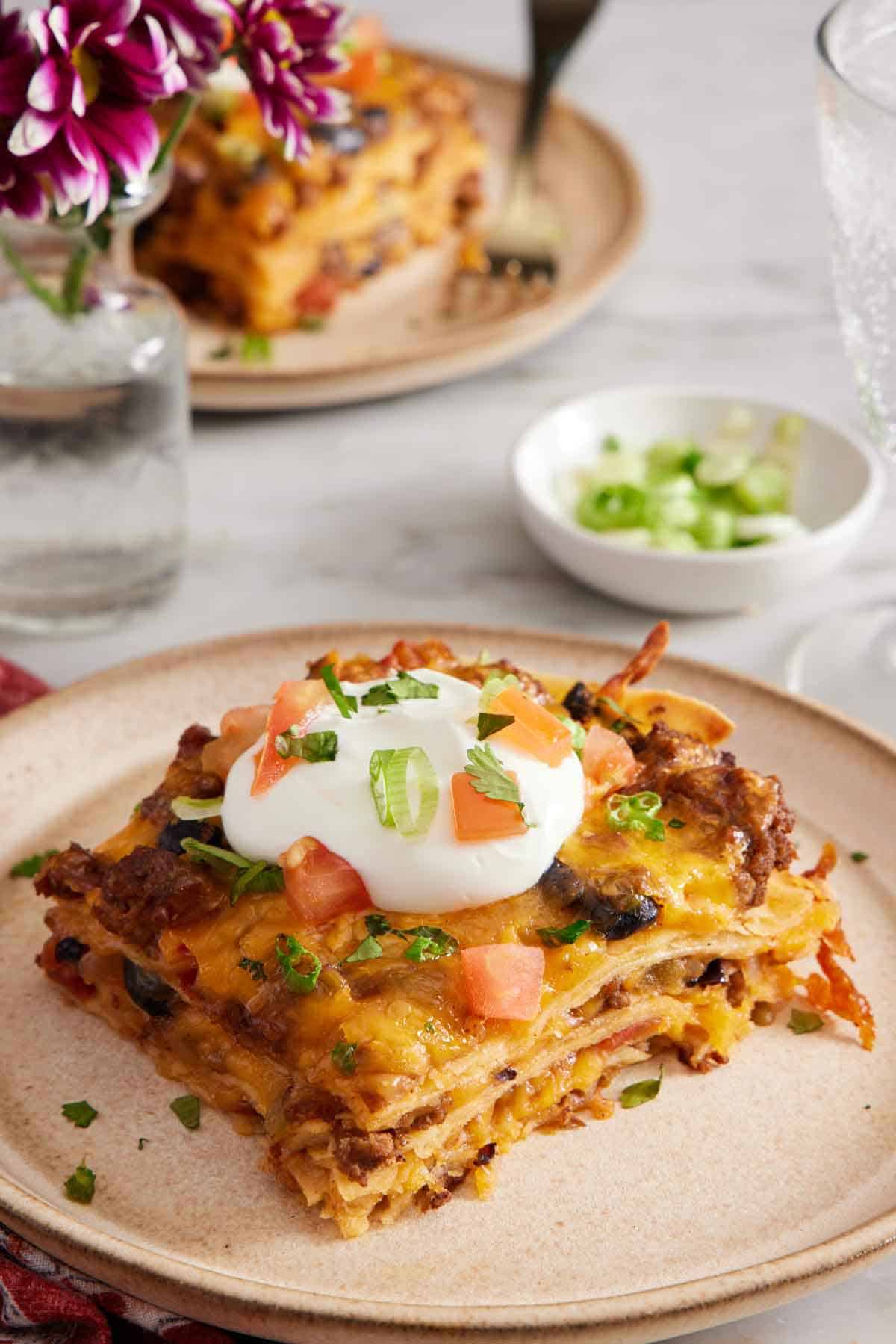 A plate with a serving of Mexican lasagna with a dollop of sour cream on top with chopped tomatoes and green onions.
