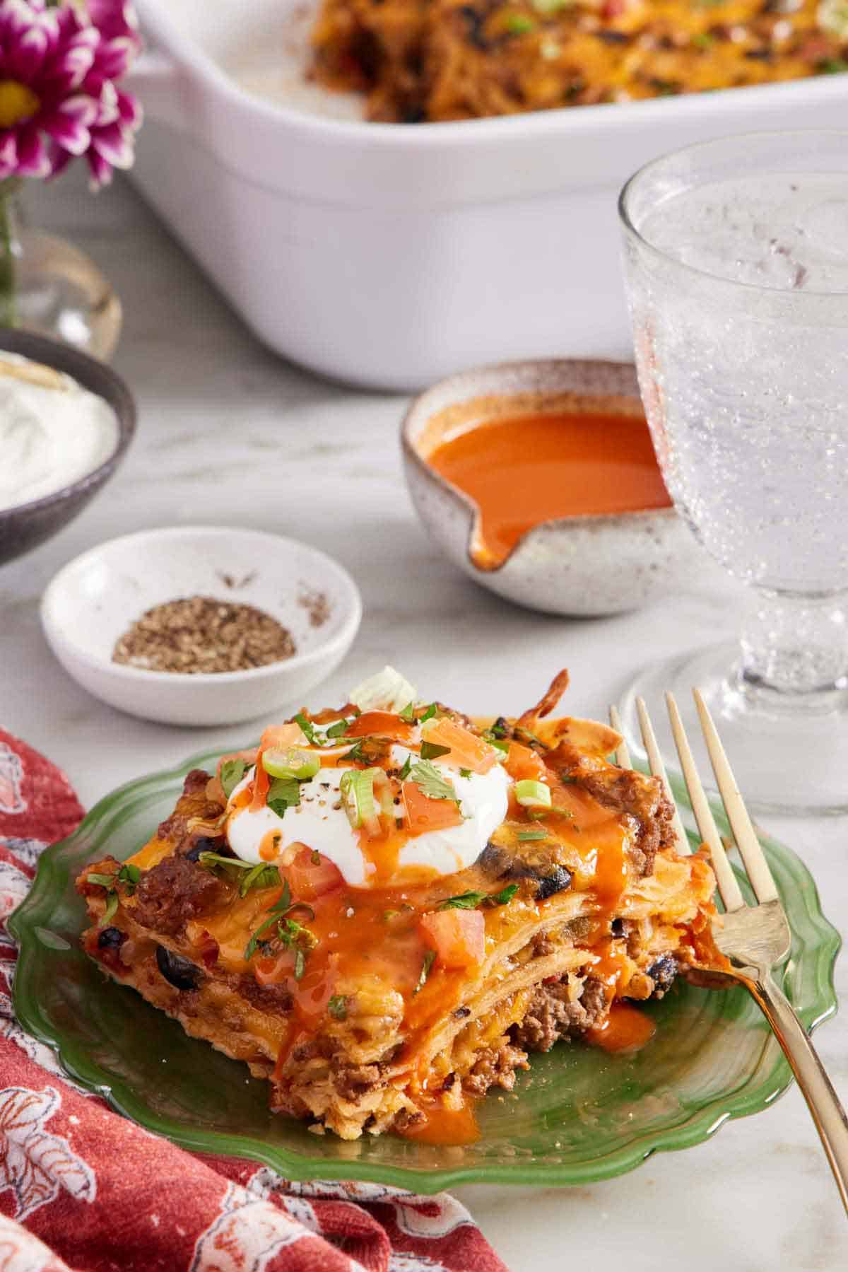 A plate with a serving of Mexican lasagna with a dollop of sour cream and hot sauce on top with chopped tomatoes and green onions. A fork on the plate and a drink in the back with toppings.