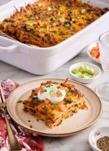 A plate with a serving of Mexican lasagna with a baking dish in the background with the rest of the lasagna. Bowls of garnishes and a drink on the side.