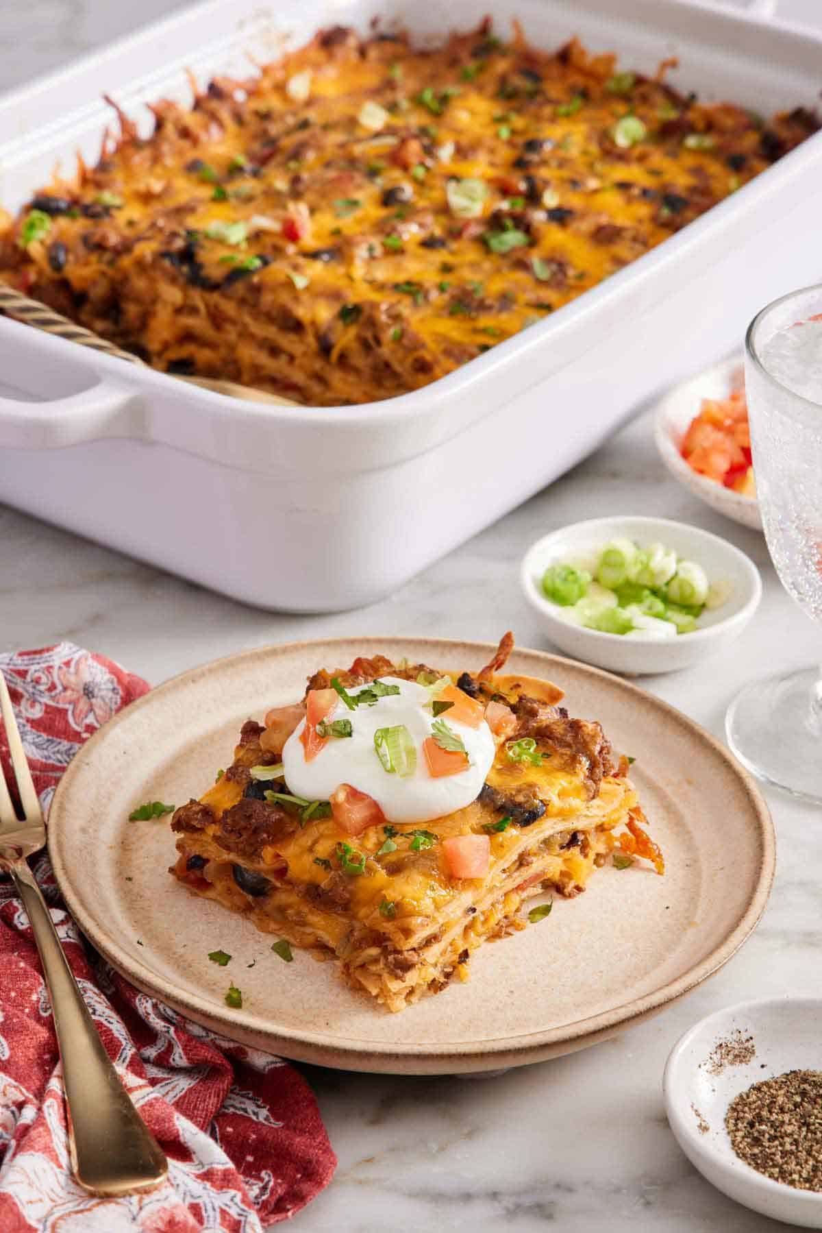 A plate with a serving of Mexican lasagna with a baking dish in the background with the rest of the lasagna. Bowls of garnishes and a drink on the side.