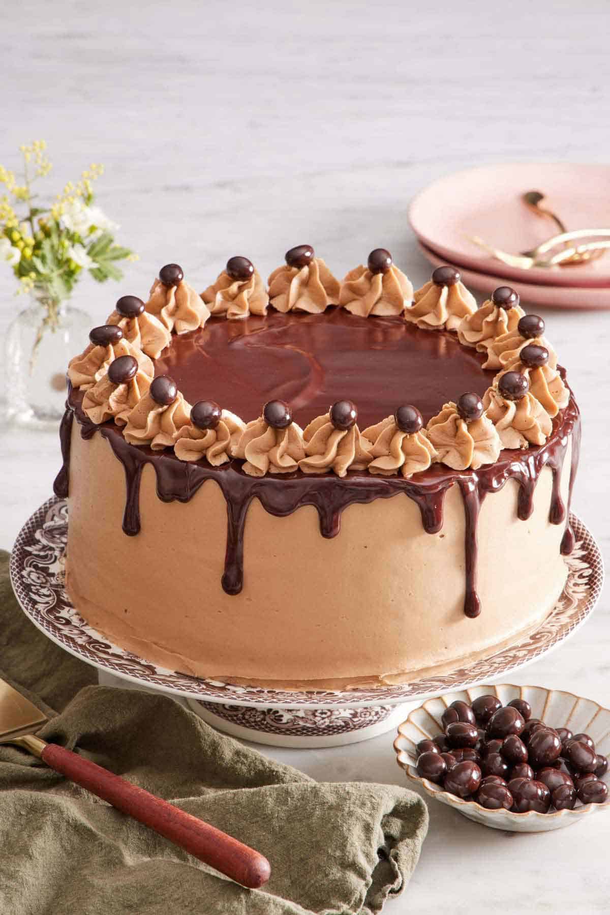 A mocha cake on a cake stand. A bowl of chocolate-covered espresso beans on the side with a cake spatula on a napkin. Flowers and a stack of plates in the background.