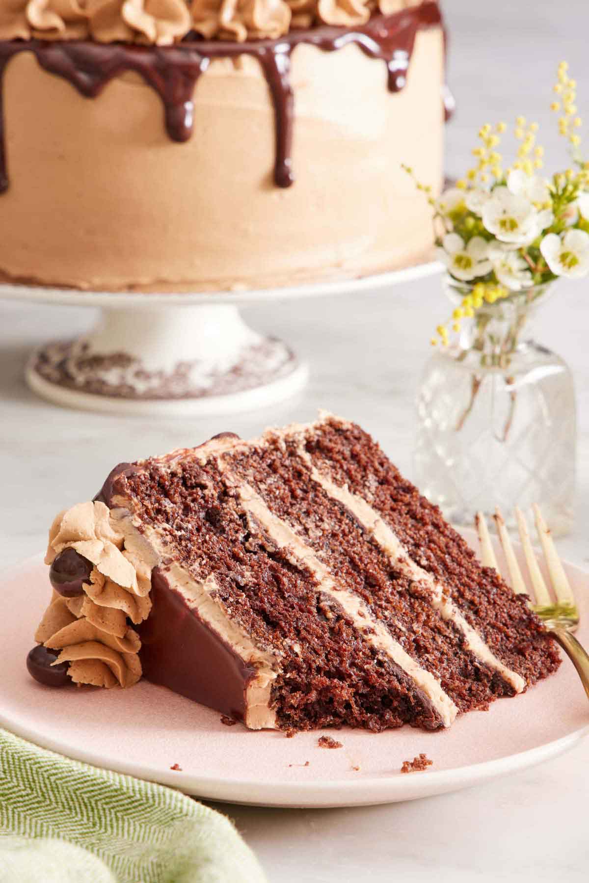 A slice of mocha cake on its side on a plate with a bite taken out. Flowers and the rest of the cake on a cake stand in the background.