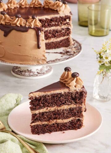 A slice of moist mocha cake on a plate with the rest of the cake on a cake stand in the background. Vase of flowers on the side.