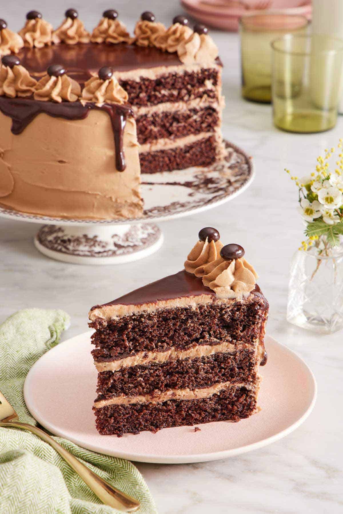 A slice of moist mocha cake on a plate with the rest of the cake on a cake stand in the background. Vase of flowers on the side.